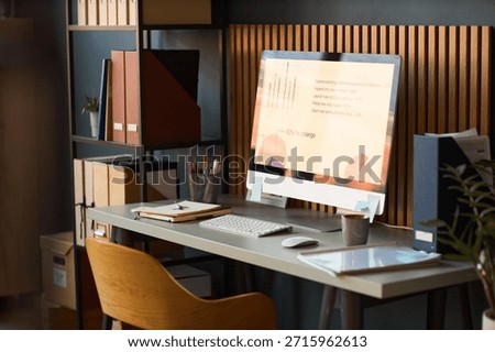 Modern workspace featuring desktop computer displaying business analytics charts and graphs on screen, organized desk with office supplies, shelving unit with folders and documents in background