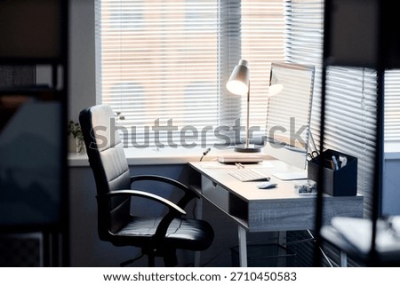 Modern workspace featuring empty office chair positioned at desk with desktop computer, keyboard, mouse, desk lamp, and office supplies near window with closed blinds