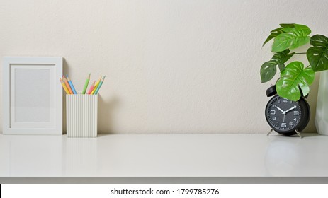 Modern Workspace With Frame Picture, Coloured Pencil, Clock And Vase On White Table In Office. Front View.