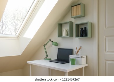 Modern workspace interior. Laptop computer, retro lamp and office accessories on a white desk. Cozy attic / loft apartment with skylight window letting the sun shine in.