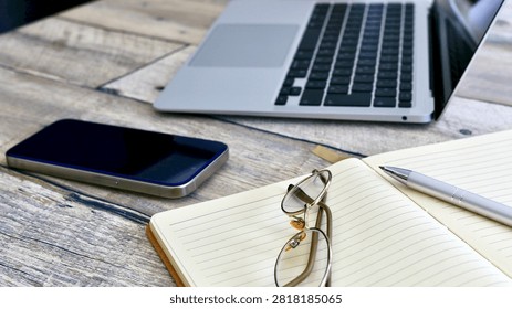 Modern workspace with an open notebook, glasses, pen, smartphone and laptop on a wooden desk, representing planning, note taking, productivity, remote work and the combination of digital and analog tools.