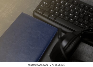 Modern workspace setup with a computer keyboard, blue leather notebook, and black headset on a wooden desk, symbolizing technology, digital communication, and remote work tools.