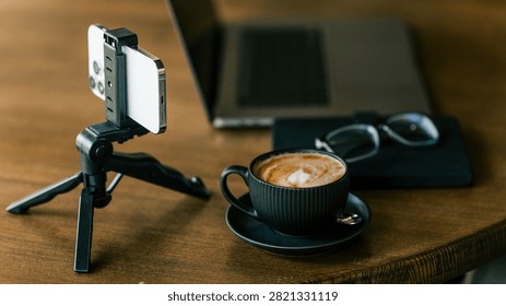A modern workspace setup featuring a smartphone on a mini tripod, a cup of latte art coffee, and a laptop, ideal for remote work or vlogging