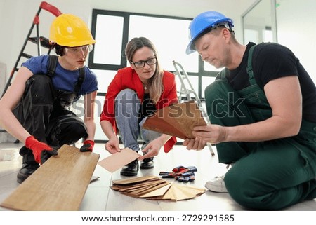 In a modern workspace, two workers and a project manager carefully examine wood samples laid out on the floor, planning for an exciting home renovation.