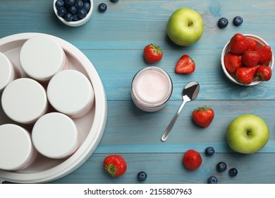 Modern yogurt maker with full jars and different fruits on light blue wooden table, flat lay