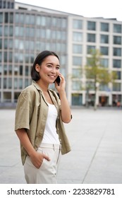 Modern young asian girl talks on mobile phone, uses telephone on city street. Woman smiling while calling someone on smartphone. - Shutterstock ID 2233829781