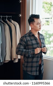 Modern Young Asian Man Putting On And Buttoning Casual Checkered Shirt While Getting Dressed At Home