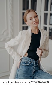 Modern young businesswoman wearing casual clothing posing indoors. Pretty female model in white suit jacket and blue jeans poses for portrait, looking at camera. Women's fashion concept. - Shutterstock ID 2220966909