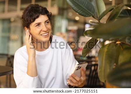 Modern young caucasian girl uses modern technology in her daily life. Brunette listens to audio message through headphones and holds smartphone sitting in cafe.
