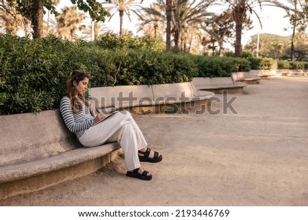 Modern young caucasian girl writes notes in notebook spending time outdoors after class. Brown-haired woman wears jacket, pants and sandals. Lifestyle concept