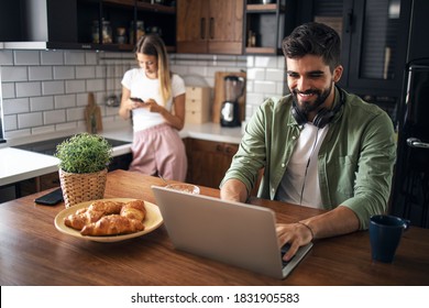 Modern young couple in the kitchen. Man is typing on laptop and smiling, women is using her cellphone in the background - Shutterstock ID 1831905583