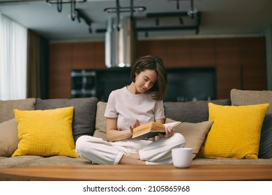 Modern young girl reading bestseller novel in paper book, enjoying weekend, relaxing on sofa at home. Self-education