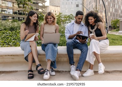 Modern Young Intercultural High School Students Work On Group Project Sitting Outdoors. Guy And Girls Use Gadgets, Notebooks. Concept Of Learning