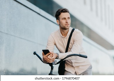 Modern young man, environmentally friendly transport and road to work. Attractive serious businessman on bicycle with bag, holds smartphone and looks away on street - Powered by Shutterstock