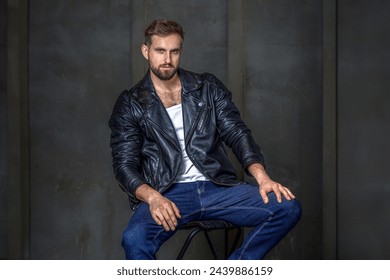 Modern young man with a stylish hairstyle, wearing a cool black leather jacket and sitting on a chair on a dark background.