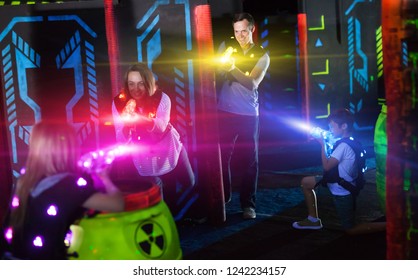 Modern young parents and children playing laser tag in dark labyrinth in bright beams of laser pistols 