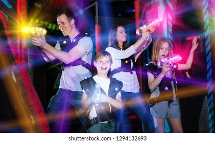 Modern young parents and children playing laser tag in dark labyrinth in bright beams of laser pistols 