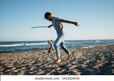 Modern Young Stylish Hipster Millennial Man Plays With A Cute Basenji Dog On The Beach At Holidays. Happy Dog Owner Enjoys Walk A Pet 