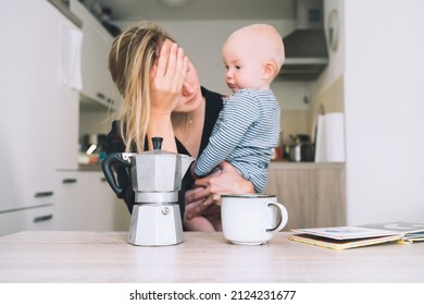 Modern Young Tired Mom And Little Child After Sleepless Night. Exhausted Woman With Baby Is Sitting With Coffee In Kitchen. Life Of Working Mother With Baby. Postpartum Depression On Maternity Leave.