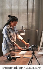 Modern Young Woman Wearing Casual Outfit Standing At Table In Loft Home Office Room Setting And Turning On Microphone To Start Vlogging