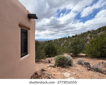 A modest adobe structure or house is built on a mountain slope. The hills are covered in pinon pines. In the distance the Sangre de Cristo mountains and the city of Santa Fe are visible. NM 2026