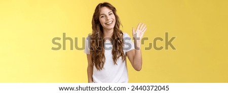 Modest cute friendly outgoing attractive european woman long curly haircut waving palm sociable conversation introduce herself smiling broadly say hi hello greeting coworker yellow background.