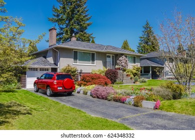 Modest Residential House With Red Car Parked On Driveway In Front. Family House With Blossoming Flowers On The Front Yard