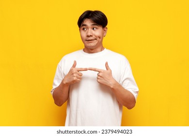 modest young asian guy in white t-shirt feeling shy on yellow isolated background, confused korean man feeling embarrassed and feeling uncomfortable