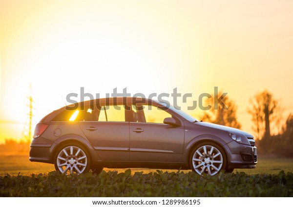 Modified image of a fictional non existent car.\
Gray car parked in countryside on blurred rural landscape and\
orange sky at sunset copy space background. Transportation,\
traveling concept.