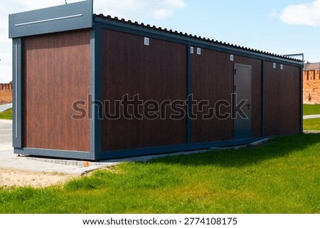 A modular building stands in an open field with grass surrounding it. The structure features a modern design with a wooden exterior and a single door, set against a clear sky.