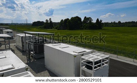 Modular containerized battery energy storage system providing grid scale power management and stability, featuring cooling units beneath a blue sky with distant power lines and green fields