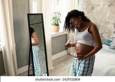 Moisturising Skin. Cheerful Pregnant Woman Taking Care Of Her Belly Skin Creaming It In Front Of The Mirror.