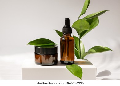 Moisturizer cream jar, serum in glass bottle on white podium with green leaves with copy space. Set for skin and body care beauty products. unbranded package, showcase.
