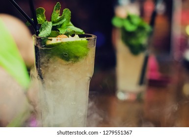 Mojito Classic Cocktails On Bar At Puff Of Smoke. Close-up Cold Cocktail With Crushed Ice, Mint And Slice Of Lime With Burning Sugar Cube On Glas. Shaker And Jigger Barmen Equipment At Background