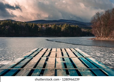 Mole (pier) on the lake. Wooden bridge in forest in winter time with blue frozen lake. Lake for fishing with pier. Dark lake (ice) with hills - Powered by Shutterstock