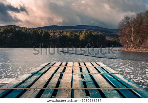 Mole (pier) on the lake. Wooden bridge in forest in\
winter time with blue frozen lake. Lake for fishing with pier. Dark\
lake (ice) with hills