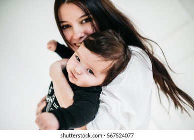 Mom And Baby. Beautiful Young Mother In A White Shirt Smiling And Holding Dark Hair Baby In Front Of White Background.