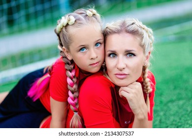 Mom And Baby Are Lying On The Grass On The Football Field Near The Gate And Look Straight.