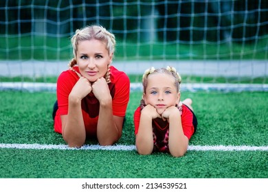 Mom And Baby Are Lying On The Grass On The Football Field Near The Gate And Look Straight.