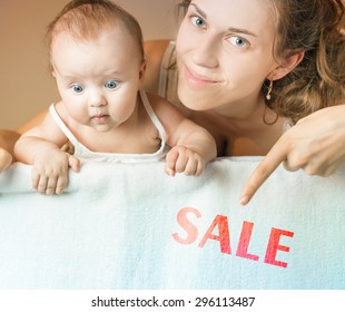 Mom And Baby Lying On White Blanket. Advertising Banner Sign - Mom Is Pointing Down On Text With Word Discount. Child Stares Down At Inscription. Mother Smiling And Looking At Camera