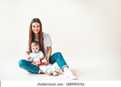 Mom And Baby Are Sitting On The Floor And Holding A Gift Box. Mom And Little Daughter Smile And Play, Beige Background With Place For Text For Mother's Day.