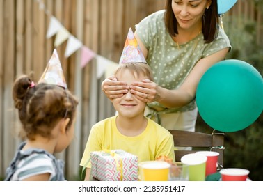 Mom from behind closes eyes her son with hands. Cute funny nine year old boy celebrating his birthday with family and friends with homemade baked cake in a backyard. Birthday party - Powered by Shutterstock