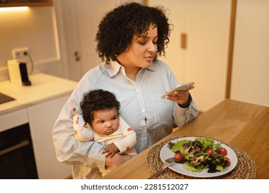 Mom blog. Hispanic curly young lady plus size mother sitting at kitchen table, holding baby toddle on her lap while have lunch, using smartphone, mommy blogger taking photo of her fresh healthy salad - Powered by Shutterstock