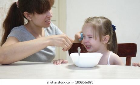 Mom Calms A Capricious Child Sitting At The Table. Parent Wipes Kid Drool And Snot With A Napkin. Teamwork. Baby Doesn T Want To Eat Porridge.