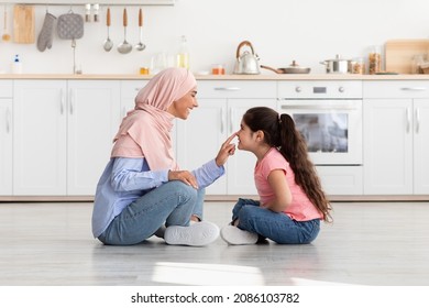 Mom And Child. Happy Loving Muslim Mother And Little Daughter Bonding In Kitchen At Home, Islamic Mother In Hijab Playfully Touching Her Kid's Nose While Sitting On Floor Together, Side View