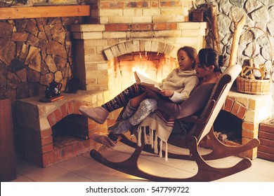 Mom With Child Reading Book And Relaxing By The Fire Place Some Cold Evening, Winter Weekends, Cozy Scene