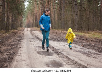 Mom And Child Running Along The Forest Road After Rain In Raincoats Together