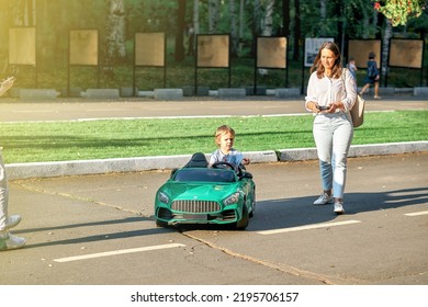 Mom Controls Children Car With Remote At Walk With Toddler Child In Public Park. Toddler Boy Sits In Green Car Holding Steering Wheel Looking Aside