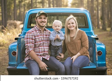 Mom, Dad And Baby Toddler Son Family Sitting On A Vintage Blue Pick-up Truck Bed And The Little One Is Clapping