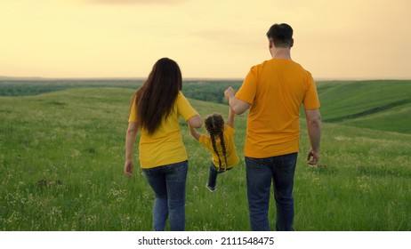 Mom, dad and daughter are playing on green grass in field, happy child is holding hands of parents and jumping. Happy family runs in park holding hands in spring, summer. Teamwork. Family weekend